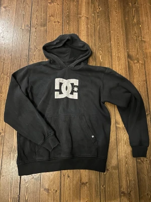 Svart DC hoodie med logga - Säljer en svart hoodie från DC med stor vit logga på bröstet. Klassisk känguruficka framtill och ribbade muddar vid ärmar och nederkant. Hoodien har en rymlig huva och är gjord i mjukt sweatshirtmaterial, perfekt för en avslappnad streetwear-look.