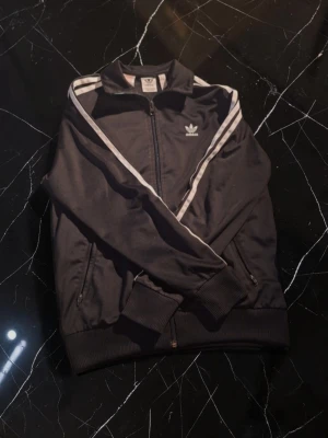 Svart Adidas track jacket - Klassisk svart Adidas track jacket med vita ränder längs ärmarna och dragkedja framtill. Jackan har ribbade muddar vid ärmslut och nederkant samt två fickor med dragkedja. Perfekt för en sportig och avslappnad stil.