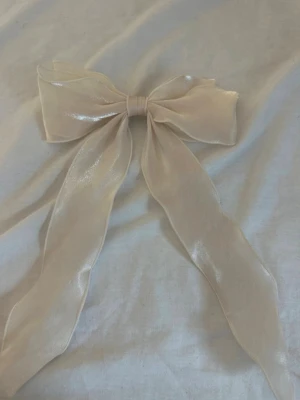 Stor cremefärgad hårrosett - Säljer en stor cremefärgad hårrosett i skimrande organza med metallspänne på baksidan. Rosetten har långa band och ger en romantisk vibe till din frisyr. Perfekt för att piffa upp håret med en söt detalj.