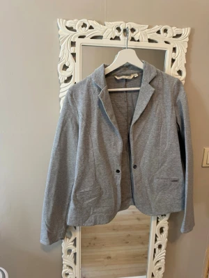 Grå kavaj med broderad detalj - Stilren grå kavaj i mjukt tyg med två knappar framtill och fickor. På ena ärmen finns en broderad svart fe-detalj som ger en unik touch. Perfekt för dig som vill ha en avslappnad men ändå snygg look.