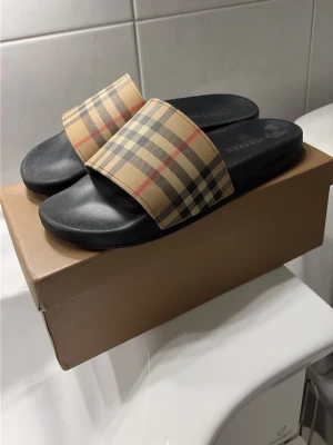 Burberry tofflor  med rutigt band - Snygga sandaler från Burberry med klassiskt beige rutigt band och svart sula. Märket är präglat i fotbädden. Perfekta för sommaren och lätta att matcha med olika outfits. Tillverkade i syntetmaterial för både komfort och stil.