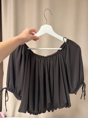 Brun croppad topp med knyt och volang - Säljer en svart offshoulder topp med korta puffärmar och knyt i sidorna. Toppen har en lös passform och resår vid halsringning och nederkant för en snygg, avslappnad look. Perfekt att styla med höga jeans eller kjol.