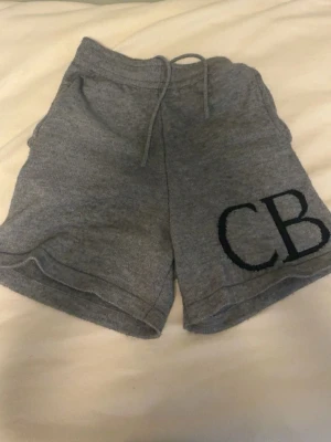 Grå mjukisshorts med snörning - Mysiga grå shorts i mjukt material med snörning i midjan och två bakfickor. På ena benet finns en stor broderad CB-logga. Perfekta för chill dagar eller träning.