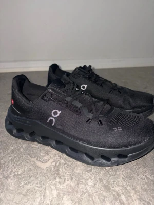 Svarta On cloud Running sneakers - Säljer ett par helsvarta On Running sneakers med ventilerande mesh-ovandel och unika, hålade sulor för extra komfort. Skorna har diskreta loggor på sidan och tungan samt svarta snören. Perfekta för dig som gillar stilrena och sportiga sneakers.
