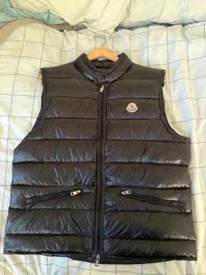 Svart dunväst från Moncler - Snygg svart dunväst från Moncler med glansig finish och klassisk logga på bröstet. Västen har hög krage med knapp, två dragkedjefickor framtill och quiltad design. Perfekt för lager-på-lager och streetwear-vibe.