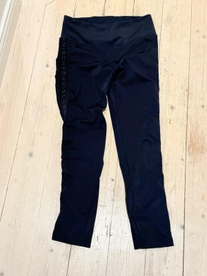 Svarta leggings med hög midja - Svarta leggings med hög midja och diskret text längs ena benet. Tillverkade i stretchigt material som sitter tajt och bekvämt. Perfekta för träning eller chill, med enkel och stilren design.