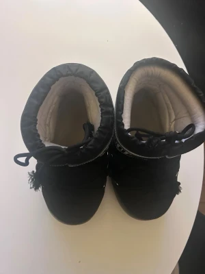 Svarta Moon Boot snowboots - Säljer ett par klassiska svarta Moon Boot snowboots med vit logga runt skaftet. Bootsens utsida är i nylon med snörning framtill och vadderad insida för extra värme. Sulan är i gummi och ger bra grepp på vintern. Perfekta för kalla dagar och snöäventyr. Strl är 39-41!!