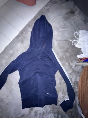 Mörkblå hoodie från GANT XS - Mörkblå hoodie från GANT i storlek XS. Klassisk modell med huva, magficka och vita ränder längs ärmarna. Tillverkad i mjukt material som känns skönt mot huden. Perfekt för en avslappnad och sportig stil.