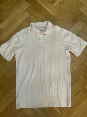 Vit ribbad polotopp från These Glory Days - Säljer en stilren vit polotopp från These Glory Days i storlek M. Toppen har ribbade vertikala ränder, klassisk krage och korta ärmar. Perfekt för dig som gillar en clean och enkel look.