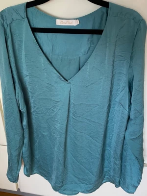 RoseBud v-ringad blus stl 46 - Säljer en ljusgrågrön blus från RoseBud i storlek 46. Blusen har v-ringning och långa ärmar, tillverkad i ett lätt och glansigt material som ger en elegant känsla. Perfekt för dig som gillar stilrena och enkla plagg. Kan variera mycket i den gröna färgen vid olika starkt solsken likt en skalbagge och bli mossgrön på kvällen.