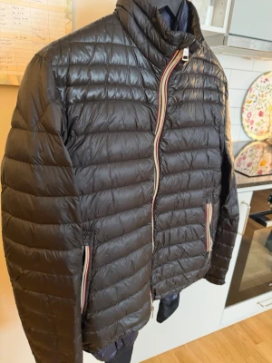 Moncler 3 - Snygg svart dunjacka med horisontella sömmar och coola randiga detaljer vid dragkedja och fickor. Jackan har hög krage och två sidofickor med dragkedja. Perfekt för kalla vinterdagar och riktigt lätt att bära.