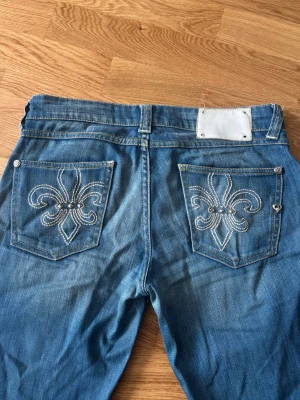 Blå bootcut jeans med broderade fickor - Säljer ett par blå bootcut jeans med snygga broderade fickor bak, dekorerade med vita sömmar i ett fleur-de-lis-mönster. Ca 40cm rakt över midjan 💕