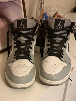Gråvita Air Jordan 1 sneakers - Säljer ett par gråvita Air Jordan 1 sneakers med svart logga och svarta skosnören. Klassisk high-top modell med perforerad tåbox och mjuk vaddering runt ankeln. Perfekta för dig som gillar streetwear och vill ha en ikonisk look.