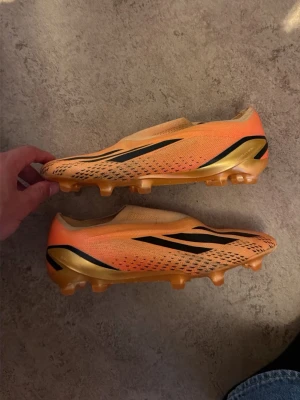 Orangea adidas fotbollsskor elite laceless - Snygga fotbollsskor i orange med svarta och guldiga detaljer. Skorna har låg profil, mönstrad ovandel och fasta dobbar i guld. Perfekta för dig som vill sticka ut på planen och ha bra grepp på konstgräs eller gräs. Knappt använda fick de av laget längesedan, men passar inte bra på mig! 