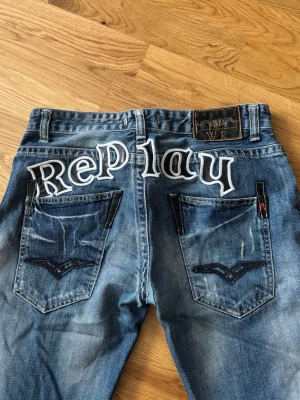 Blå Replay jeans med brodyr - Säljer ett par blå jeans från Replay med stor vit brodyrtext över bakfickorna och slitna detaljer. Ca 41cm rakt över midjan 💕