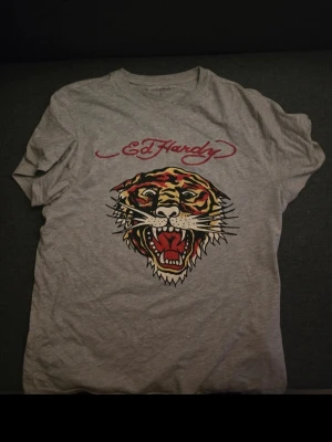 Grå Ed Hardy t-shirt med tigertryck - Säljer en grå t-shirt från Ed Hardy med ett stort tigertryck framtill och röd logga ovanför. T-shirten är kortärmad och har en klassisk passform. Perfekt för dig som gillar streetstyle och vill sticka ut med en cool design.