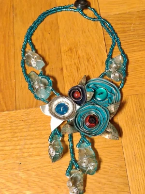 Turkost halsband med pärlor och metall - Unikt halsband med turkosa och silverfärgade pärlor, spiralformade metalltrådar och dekorativa knappar. Halsbandet har ett blommigt motiv i mitten och detaljer av glasliknande stenar och metallblad. Perfekt för dig som gillar statement-smycken och färgstarka accessoarer.