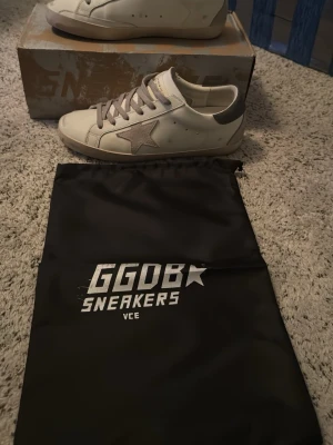 Vita GGDB Superstar sneakers - Säljer ett par vita Golden Goose Superstar sneakers med den ikoniska stjärnan på sidan och grå detaljer. Skorna har låga skaft, vita snören och en slitstark gummisula. Kommer med originalkartong och dustbag. Perfekta för dig som gillar en avslappnad men trendig stil.