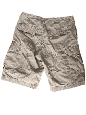 Beige cargoshorts från Carhartt - Säljer ett par beige cargoshorts från Carhartt med flera praktiska fickor och klassisk passform. Shortsen har bälteshällor och stängs med knapp och dragkedja. Perfekta för sommardagar och avslappnade outfits.Midja 50 cm Lång sida 62 cm Ben öppning 33 cm