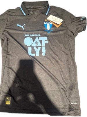 Malmö FF matchtröja Puma svart - Snygg svart Malmö FF matchtröja från Puma med ljusblå detaljer och klubbmärke på bröstet. Tryck med Oatly och Akea på fram- och baksidan. Tillverkad i funktionsmaterial Drycell som andas, perfekt för träning eller match.