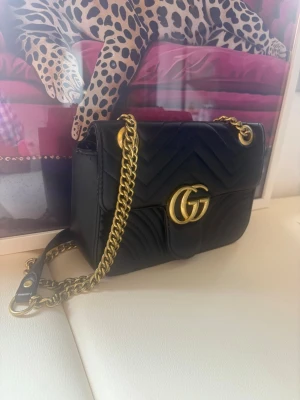 Svart  Marmont handväska - Snygg svart Marmont handväska i quiltat läder med guldfärgade detaljer och kedjerem. Väskan har GG-logga framtill och klassisk klaffstängning. Perfekt storlek för det viktigaste och ger en lyxig touch till din outfit.