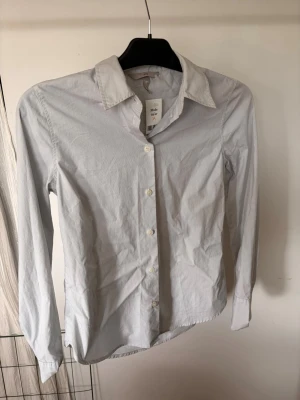 Klassisk randig skjorta med lång ärm - Stilren vit och blå randig skjorta med lång ärm och knäppning framtill. Skjortan har en klassisk krage och är tillverkad i ett tunt, vävt material som ger en clean look. Perfekt till jeans eller kostymbyxor för en enkel och snygg outfit.