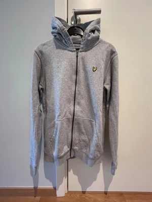 Grå zip hoodie Lyle&Scott - Säljer en grå hoodie med dragkedja och huva. Tröjan har en broderad gul logga på bröstet, två fickor framtill och ribbade muddar vid ärmar och nederkant. Tillverkad i mjukt sweatshirtmaterial, perfekt för en avslappnad stil.