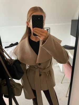 Beige kappa med stor krage från NA-KD - Trendig beige kappa från NA-KD med oversized krage och knytskärp i midjan. Kappan har två knappar framtill och raka ärmar. Tillverkad i ett mjukt material som ger en stilren och modern look.