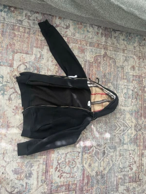 Svart hoodie från Burberry - Säljer en svart hoodie från Burberry i storlek M. Hoodien har dragkedja, fickor framtill och en ikonisk beige-rutig huva med röda och grå detaljer. Helt fel fri!                                                             Pris kan diskuteras 