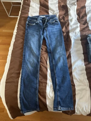 Blå bootcut jeans med slitningar - Säljer ett par blå bootcut jeans med lätt slitna detaljer och klassiska fem fickor. Jeansen har normal midja och är gjorda i denim med lite stretch för skön passform. Perfekta till sneakers eller boots. Märke pepe jeans storlek 27:34, lågmidjade.
