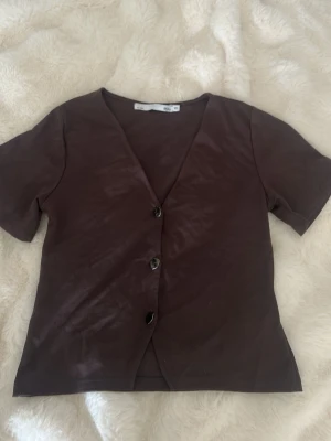 Brun kortärmad topp med knappar - Snygg brun topp med korta ärmar och v-ringning. Toppen har tre knappar framtill och är i ett mjukt material som känns skönt mot huden. Perfekt att styla med jeans eller kjol för en trendig look. Aldrig använd men haft i garderoben 