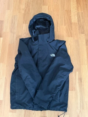 Svart vindjacka från The North Face - Svart vindjacka med huva från The North Face. Jackan har dragkedja framtill, två sidofickor med dragkedja och justerbar dragsko i nederkant. Tillverkad i ett lätt och vattenavvisande material, perfekt för blåsiga dagar.
