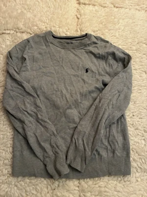 Grå stickad tröja från Polo Ralph Lauren - Snygg grå stickad tröja från Polo Ralph Lauren med klassisk rund halsringning och diskret logga broderad på bröstet. Tröjan har långa ärmar och ribbade muddar vid ärmslut och nederkant. Perfekt för lager-på-lager och enkel att matcha.