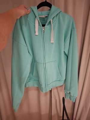 Turkos zip hoodie med huva - Snygg turkos hoodie med dragkedja och vita snören i huvan. Hoodien har känguruficka framtill och ribbade muddar vid ärmar och nederkant. Perfekt för en avslappnad och chill stil. Materialet känns mjukt och bekvämt.
