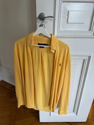 Gul långärmad skjorta med logga - Snygg gul skjorta med lång ärm och klassisk krage. Skjortan har en liten broderad logga på bröstet och knappar framtill. Perfekt för dig som vill ha en färgklick i garderoben.