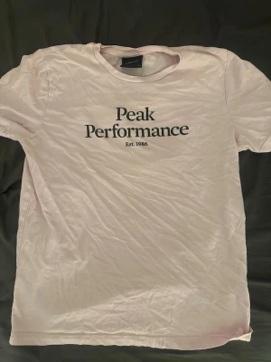 Ljusrosa Peak Performance t-shirt - Säljer en ljusrosa t-shirt från Peak Performance med svart tryck på bröstet där det står 'Peak Performance Est. 1986'. Klassisk rund hals och korta ärmar. Perfekt till jeans eller shorts för en avslappnad look. Är i storlek 150 men passar xs 