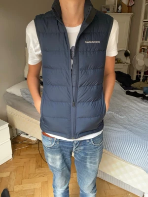 Mörkblå dunväst Peak Performance - Snygg mörkblå dunväst från Peak Performance med hög krage och diskret logga på bröstet. Västen har quiltad design och dragkedja framtill. Perfekt att slänga över en hoodie eller t-shirt för extra värme och stil.
