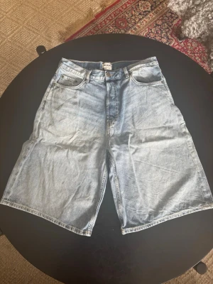 Ljusblåa bermudashorts med baggy fit - Säljer ett par ljusblåa jeansshorts i loose fit med mid till low waist. Klassisk femficksmodell och dragkedja med knapp. Perfekta för sommaren och ger en avslappnad vibe till din outfit. Storlek 30 i Weekday sizing (30-inch waist), funkar som S/M.
