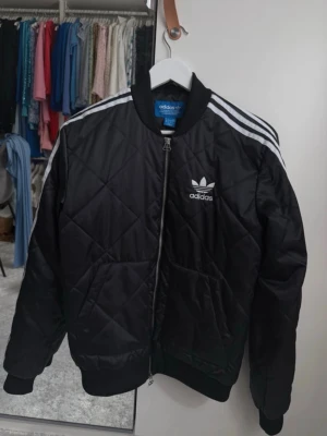 Svart bomberjacka från Adidas - Snygg svart bomberjacka från Adidas med klassiska vita ränder på axlarna och broderad logga på bröstet. Jackan har quiltat mönster, ribbade muddar och dragkedja framtill. Perfekt för dig som gillar sportig streetstyle.