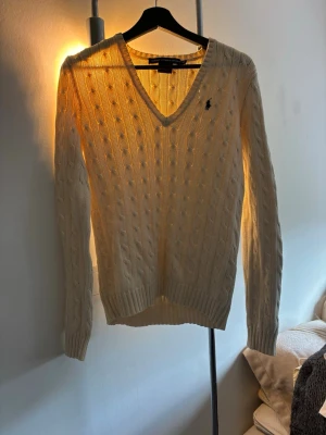 Vit kabelstickad tröja Ralph Lauren - Kabelstickad vit tröja från Ralph Lauren med V-ringning och lång ärm. Tröjan har broderad logga på bröstet och ribbade muddar vid ärmslut och nederkant. Klassisk och stilren modell som passar till allt. Storlek m men passar S 