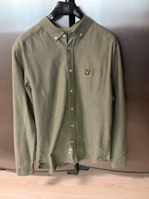 Lyle&Scott skjorta - Olivgrön skjorta i bomull med klassisk krage och knäppning framtill. Bröstficka med broderad gul logga. Långärmad och enkel, passar perfekt till jeans eller chinos.