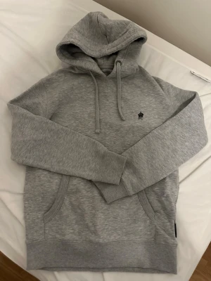 French connection grå hoodie  - Snygg grå hoodie med huva och dragsnören. Har en liten svart broderad logga på bröstet och praktisk magficka framtill. Tillverkad i mjukt sweatshirtmaterial och har ribbade muddar vid ärmar och nederkant. Perfekt för en chill och avslappnad stil.