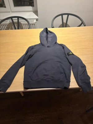Mörkblå stone Island hoodie  - Säljer en mörkblå hoodie med huva och känguruficka framtill. På vänster ärm sitter en svart patch med gul och grön logga. Hoodien har ribbade muddar vid ärmslut och nederkant. Perfekt för dig som gillar streetwear och enkel stil. Storlek 156