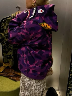 Bape a bathing ape hoodie - Säljer en unik hoodie i lila och rosa camomönster med hajmotiv på huvan och dragkedja framtill. Hoodien har ribbade muddar och fickor på magen. Perfekt för dig som vill sticka ut med streetwear-vibe.