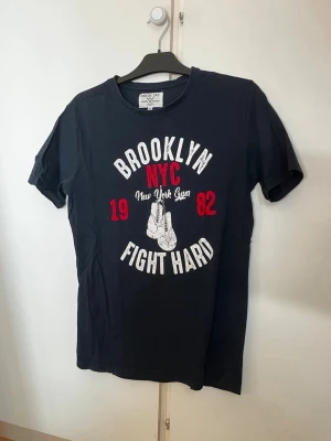 Svart Brooklyn NYC t-shirt - Cool svart t-shirt med tryck i vitt och rött där det står 'Brooklyn NYC Fight Hard' och en illustration av boxningshandskar. T-shirten har rund hals och korta ärmar. Perfekt för dig som gillar streetwear och sportig stil.