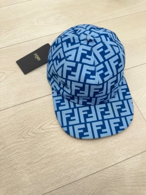 Blå Fendi keps med FF-mönster - Snygg blå keps från Fendi med ikoniskt FF-mönster i olika blå nyanser. Kepsen har böjd skärm och justerbar baksida. Tillverkad i ett slitstarkt tyg som ger en cool och trendig look. Perfekt accessoar för att lyfta din outfit.