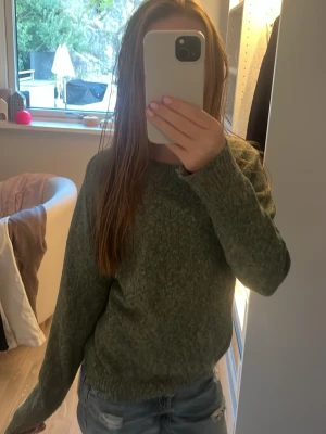 Grön stickad tröja med rund hals - Mysig grön stickad tröja med rund halsringning och långa ärmar. Tröjan har ribbstickade muddar vid ärmslut och nederkant. Perfekt för kyligare dagar och enkel att matcha med jeans eller kjol.