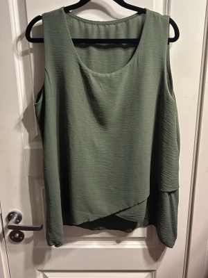 Olivgrön ärmlös topp med omlott - Säljer en olivgrön ärmlös topp med rund halsringning och snygg omlottdetalj framtill. Tyget är lätt och har en mjukt strukturerad yta som ger en avslappnad känsla. Perfekt att styla med jeans eller kjol för en trendig look.