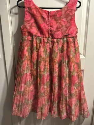 Blommig rosa klänning med plisserad kjol - Söt ärmlös klänning med blommigt mönster i rosa, grönt och orange. Överdelen är figursydd och kjolen är plisserad för extra volym. Dragkedja bak. Perfekt för dig som gillar romantisk och färgglad stil.