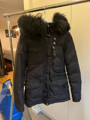 Svart parkas med fuskpäls - Snygg svart parkas med stor huva och fluffig fuskpäls. Jackan har flera fickor framtill, dragkedja och knappar samt justerbar midja. Perfekt för kalla dagar och riktigt varm. Klassisk modell som funkar till det mesta.